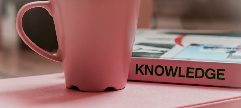 Pink cup on the pink table, behind there is a pink book titled Knowledge