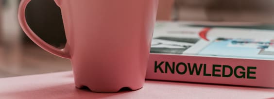 Pink cup on the pink table, behind there is a pink book titled Knowledge